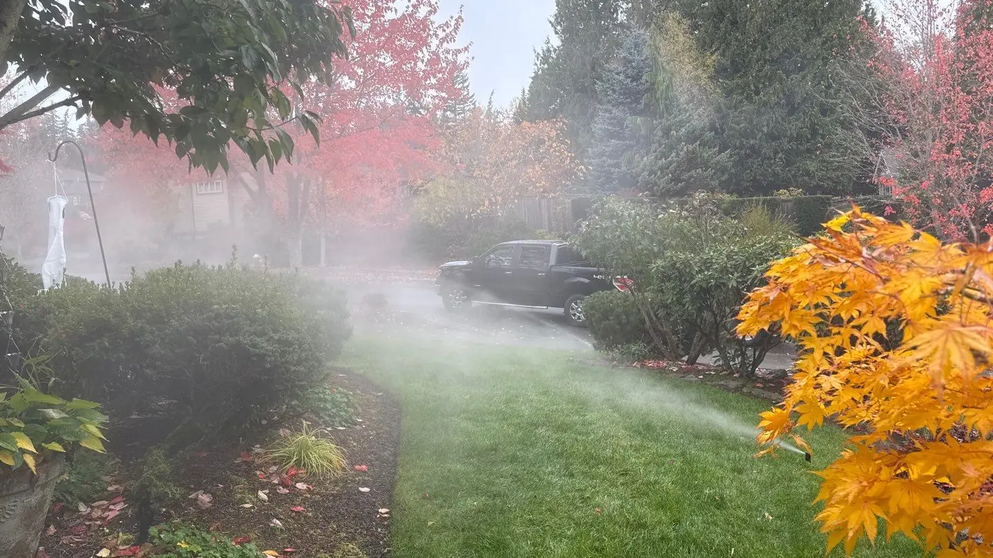 Mist drifting over a front lawn as a sprinkler zone is blown out, with a black pickup parked beyond it